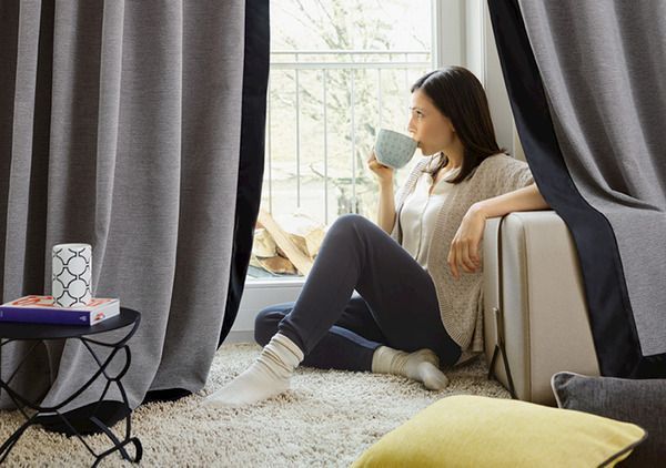 Eine Frau sitzt am Fenster zwischen Vorhängen und trinkt dabei aus einer Tasse.